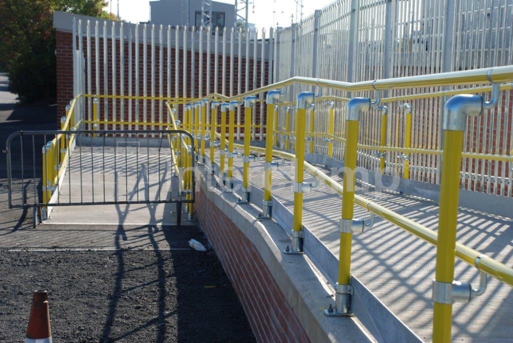 Bright yellow handrail tube and Interclamp Key Clamp DDA Assist fittings forming a safe and compliant disabled access ramp at a train station, promoting inclusivity.